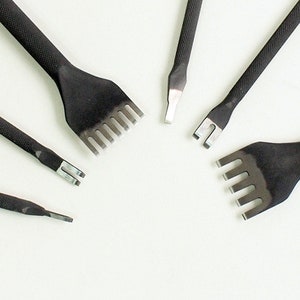 May include: A collection of black leather working tools, including various sizes of stitching chisels and a groover. The tools are arranged on a white surface, showcasing their sharp, metal tips and textured handles. These tools are used for leathercraft.