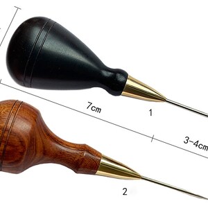 May include: Two leather awls with wooden handles. The awl on the left has a black handle and is 7 inches long. The awl on the right has a brown handle and is 7 inches long. Both awls have a gold metal tip.