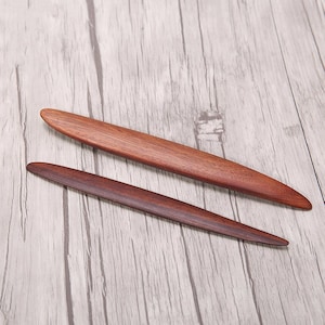 May include: Two wooden hair sticks, one dark brown and one light brown, on a light wood surface.