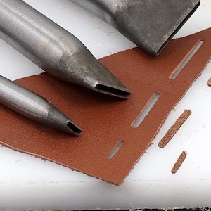Puede incluir: Tres punzones de cuero de metal de diferentes tamaños, con un trozo de cuero marrón que muestra los agujeros perforados por las herramientas.