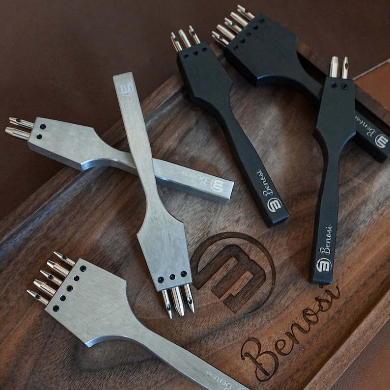 May include: Four leather hole punch tools on a wooden surface. The tools are made of metal and have different numbers of prongs. The tools are labeled "Benossi" and have a logo of a stylized "B".