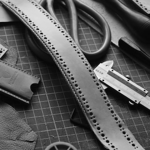 May include: A black and white image of leatherworking tools and materials. A leather strap with a row of holes is laid out on a gridded cutting mat. A ruler, a pair of scissors, a metal caliper, and a leather punch are also visible.