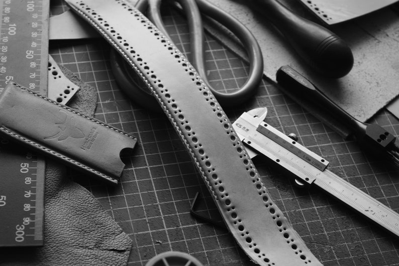 May include: A black and white image of leatherworking tools and materials. A leather strap with a row of holes is laid out on a gridded cutting mat. A ruler, a pair of scissors, a metal caliper, and a leather punch are also visible.