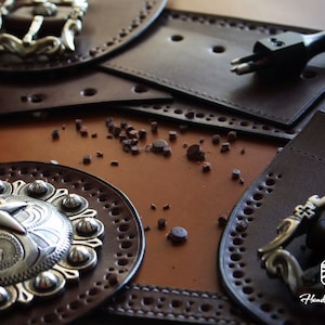 May include: Close-up of a leather craft project with two silver buckles, a leather punch tool, and a brown leather background. The leather pieces are decorated with stitching and holes.