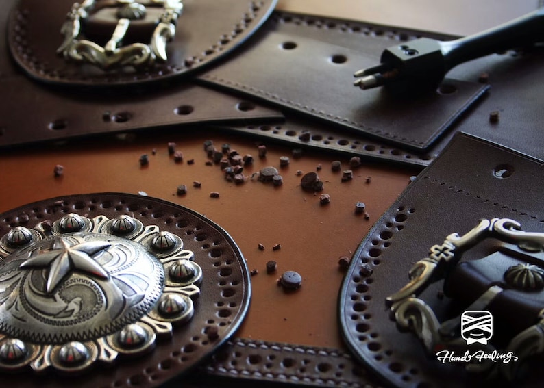 May include: Close-up of a leather craft project with two silver buckles, a leather punch tool, and a brown leather background. The leather pieces are decorated with stitching and holes.
