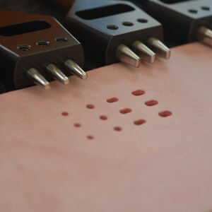 Pu&ograve; includere: Primo piano di quattro strumenti di punzonatura in pelle nera con punte in metallo argentato. Gli strumenti sono disposti su una superficie in pelle rosa chiaro con un motivo di piccoli fori punzonati.