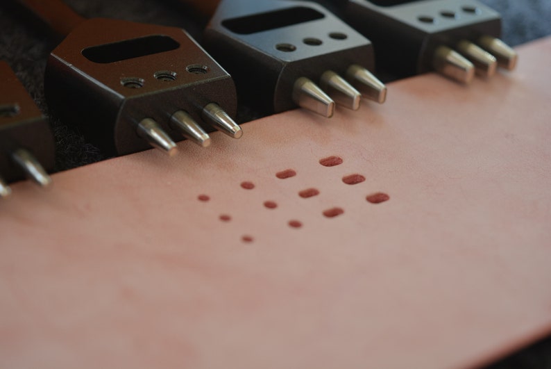 Pu&ograve; includere: Primo piano di quattro strumenti di punzonatura in pelle nera con punte in metallo argentato. Gli strumenti sono disposti su una superficie in pelle rosa chiaro con un motivo di piccoli fori punzonati.