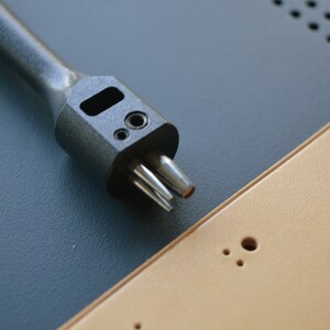 May include: A black leather punch tool with multiple prongs, resting on a tan leather surface with multiple holes.