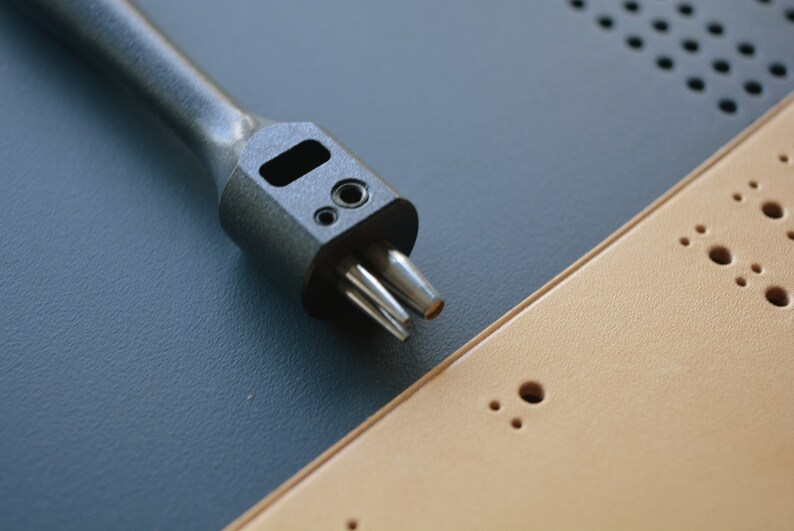 May include: A black leather punch tool with multiple prongs, resting on a tan leather surface with multiple holes.