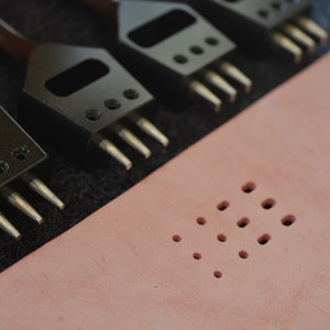Pu&ograve; includere: Quattro strumenti di punzonatura in pelle nera con punte metalliche su una superficie in tessuto nero. Un pezzo di pelle rosa con una griglia di fori punzonati.