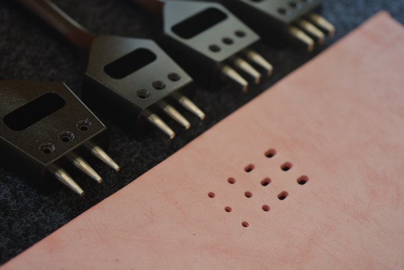 Pu&ograve; includere: Quattro strumenti di punzonatura in pelle nera con punte metalliche su una superficie in tessuto nero. Un pezzo di pelle rosa con una griglia di fori punzonati.