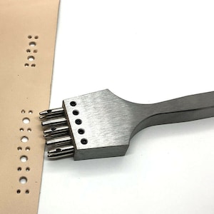 May include: A silver leather punch tool with a handle and multiple punch heads. The tool is being used to punch holes in a piece of beige leather.