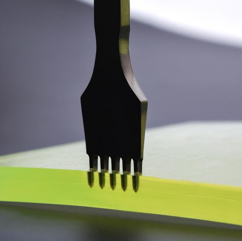 May include: A black tool with five prongs is pressing into a sheet of green material. The tool is used for making holes in leather or other materials.