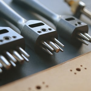 May include: Three metal leather hole punches with different sized tips. The punches are on a brown leather surface with holes punched in it.
