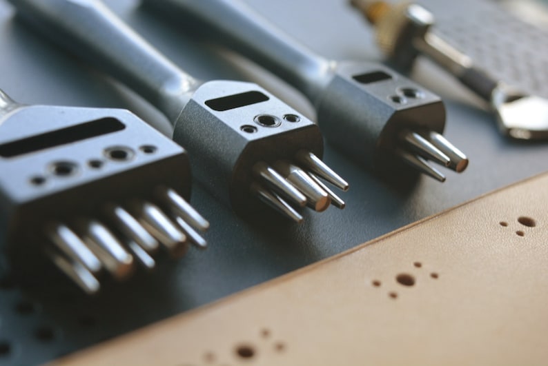 May include: Three metal leather hole punches with different sized tips. The punches are on a brown leather surface with holes punched in it.