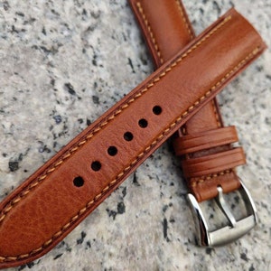 Pu&ograve; includere: Un cinturino da orologio in pelle marrone con un bordo cucito e una fibbia argentata.