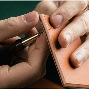 May include: A person using a leather stitching tool to sew two pieces of brown leather together.