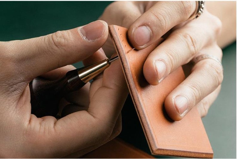 May include: A person using a leather stitching tool to sew two pieces of brown leather together.