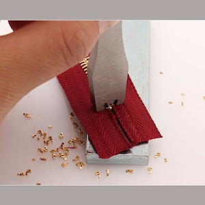 Op de afbeelding: Een close-up van een rode rits die wordt gesloten met een metalen gereedschap. Het gereedschap wordt gebruikt om de rits tanden samen te knijpen. Er liggen kleine metalen stukjes rond de rits.