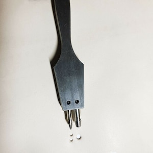 May include: A close-up of a silver leather punch tool with a wooden handle. The tool has three prongs and is used to create holes in leather.