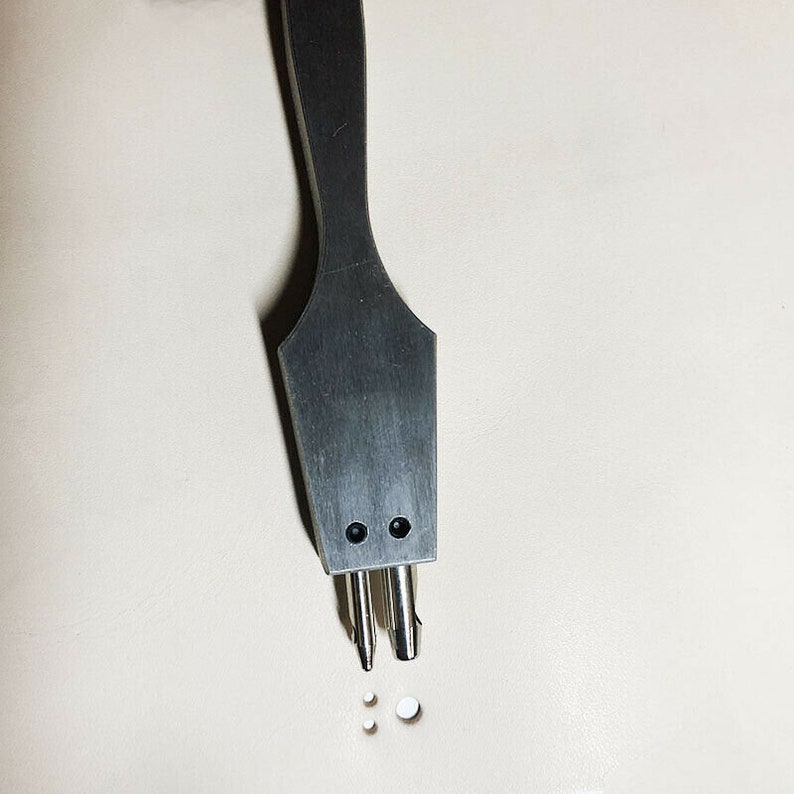 May include: A close-up of a silver leather punch tool with a wooden handle. The tool has three prongs and is used to create holes in leather.
