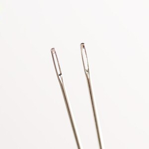 May include: Two silver sewing needles on a white background.