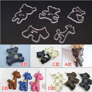 Può includere: Cinque modelli in plastica trasparente per realizzare portachiavi in pelle. I modelli sono a forma di orso, giraffa, gatto e due orsi diversi. Sotto i modelli ci sono cinque portachiavi in pelle finiti, uno per ogni design. I portachiavi sono realizzati in pelle marrone, nero, blu, rosa e giallo. I portachiavi sono etichettati con le lettere da A a E.
