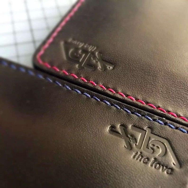 May include: Two black leather wallets with contrasting stitching. The top wallet has pink stitching and the text "Barry Bill" and the bottom wallet has blue stitching and the text "The Iowa".