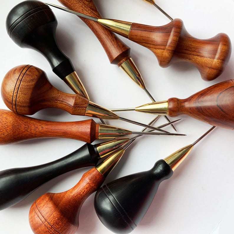 May include: A set of eight leather crafting tools with wooden handles and metal tips. The tools are arranged in a starburst pattern on a white surface.