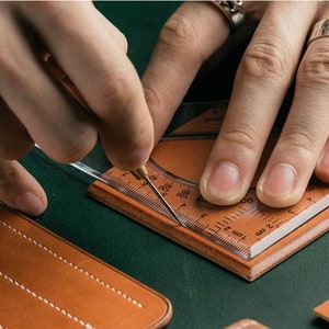 May include: A person is using a clear plastic ruler and a small awl to mark a piece of brown leather. The ruler has markings in inches and centimetres.