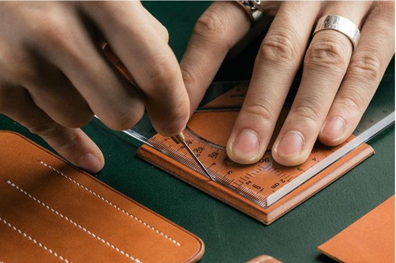 May include: A person is using a clear plastic ruler and a small awl to mark a piece of brown leather. The ruler has markings in inches and centimetres.