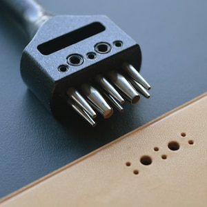 May include: A black leather punch tool with multiple sized punches. The tool is lying on a tan leather surface with holes punched in it.