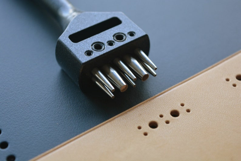 May include: A black leather punch tool with multiple sized punches. The tool is lying on a tan leather surface with holes punched in it.