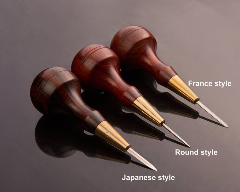 May include: Three different styles of leather awls with wooden handles and metal tips. The awls are labelled "Japanese style", "Round style", and "France style".