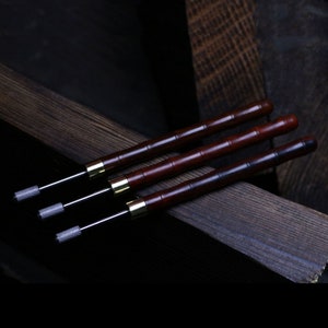 May include: Three wooden tools with metal tips. The tools have a dark brown wooden handle and a gold band near the tip. The tools are arranged on a dark wooden surface.