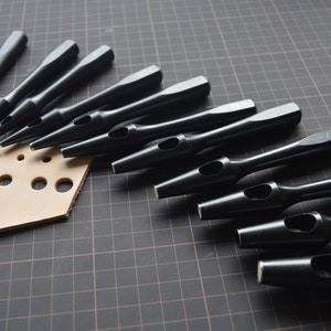 Könnte beinhalten: Ein Satz von zehn schwarzen Lederlochstempeln in verschiedenen Größen. Die Stempel sind in einem Halbkreis auf einer schwarzen Gitteroberfläche angeordnet. Ein Stück braunes Leder mit gelochten Löchern befindet sich in der linken unteren Ecke des Bildes.