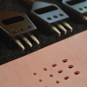 Pu&ograve; includere: Primo piano di quattro strumenti per bucare il cuoio con punte metalliche. Gli strumenti sono disposti in fila su una superficie nera. Un pezzo di pelle rosa con pi&ugrave; fori si trova sullo sfondo.