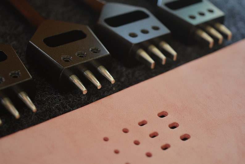 Pu&ograve; includere: Primo piano di quattro strumenti per bucare il cuoio con punte metalliche. Gli strumenti sono disposti in fila su una superficie nera. Un pezzo di pelle rosa con pi&ugrave; fori si trova sullo sfondo.