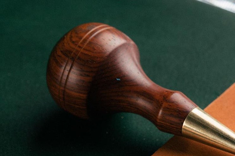 May include: A close-up of a wooden leather crafting tool with a brass tip. The tool is made of dark wood with a grain pattern and has a rounded handle.