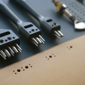 May include: A set of four leather hole punches with different sized tips. The punches are black and silver and are arranged on a gray surface. The punches are used to create holes in leather for stitching or other purposes.