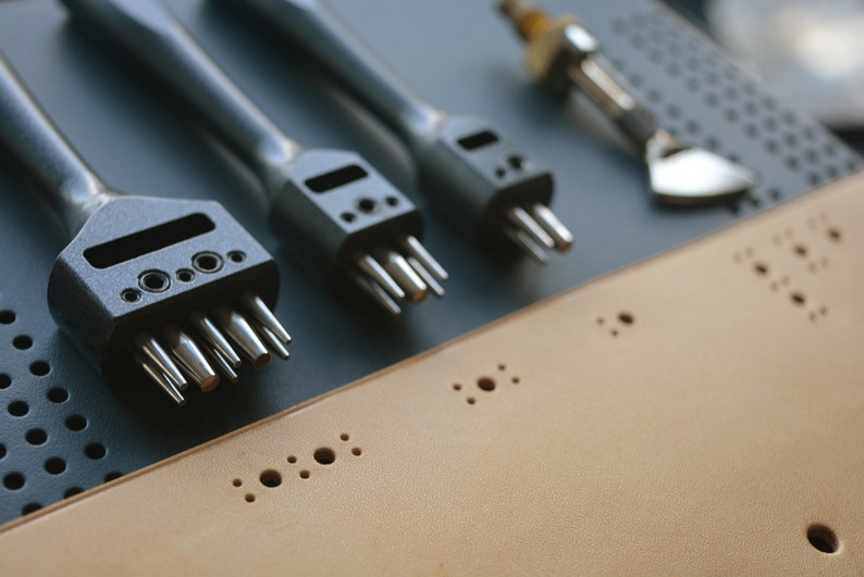 May include: A set of four leather hole punches with different sized tips. The punches are black and silver and are arranged on a gray surface. The punches are used to create holes in leather for stitching or other purposes.