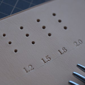 Pu&ograve; includere: Primo piano di uno strumento per la lavorazione del cuoio con una fila di fori e segni per 1,2, 1,5, 1,8 e 2,0 pollici. Lo strumento viene utilizzato per perforare il cuoio.