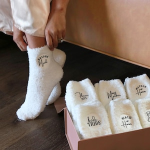 May include: White fuzzy socks with black text, including "Bridesmaid," "Flower Girl," "Bride Tribe," "Mother," "Matron of Honor," "Maid of Honor," and "Bride." The socks are in a pink box, with one being worn.