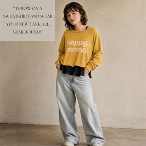 Puede incluir: Una sudadera corta de manga larga en color amarillo mostaza con el texto "happy mama" en blanco, superpuesta sobre una camiseta sin mangas negra, combinada con vaqueros anchos de lavado claro. La imagen incluye el texto "THROW ON A SWEATSHIRT AND WEAR YOUR NEW TANK ALL YEAR ROUND!"