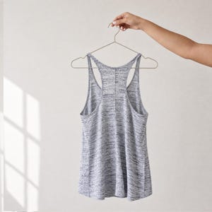 Puede incluir: Una camiseta de tirantes con espalda nadadora en color gris y blanco jaspeado colgada de una percha de metal fina. La prenda presenta un dise&ntilde;o sin mangas con cuello redondo y espalda nadadora, con un patr&oacute;n de tela texturizado y moteado.