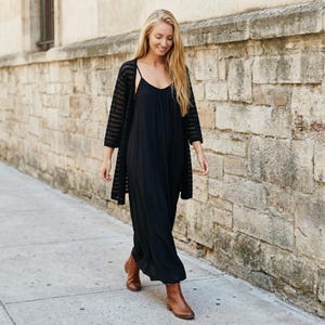 May include: A black maxi dress with thin spaghetti straps paired with a black sheer cardigan featuring horizontal stripes. The outfit is completed with brown leather ankle boots, creating a layered look with a flowing silhouette and simple design.
