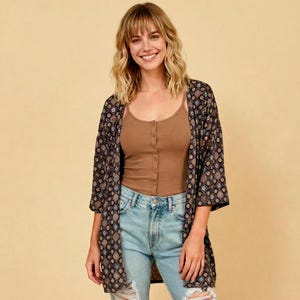 May include: A brown ribbed vest top with a button-down front, paired with light blue denim jeans and a black open-front kimono featuring a repeating geometric pattern in shades of blue, white, and tan. The kimono has wide, three-quarter length sleeves.