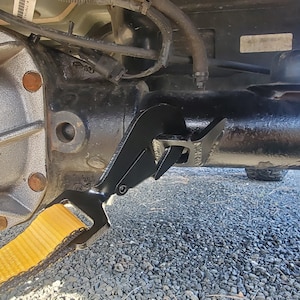 May include: A black metal strap with a yellow strap attached to it. The strap is hooked to a black metal axle. The strap is used to secure a load.