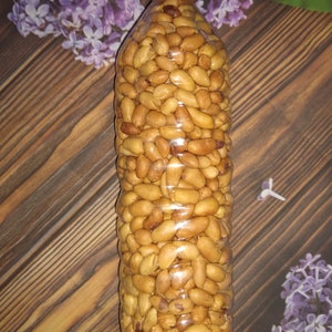 May include: A clear plastic bottle filled with roasted peanuts. The bottle has a yellow cap and is filled with golden-brown peanuts. The background features a wooden surface and purple flowers.