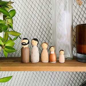 May include: A set of six wooden peg dolls, painted in various shades of brown, pink, and white. The dolls are standing on a wooden shelf with a wire mesh background.
